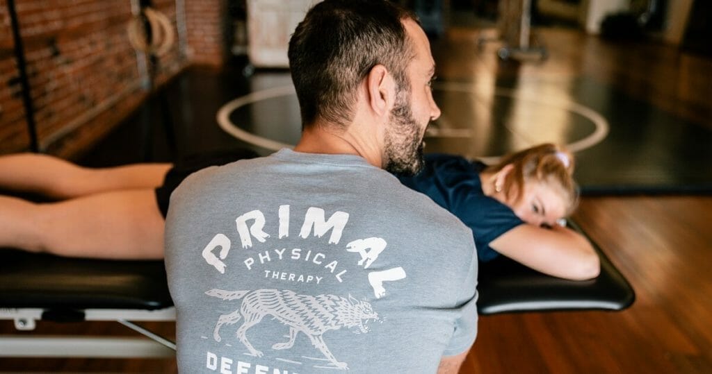 a man with primal physical therapy shirt performing a session with a woman laying on the bench