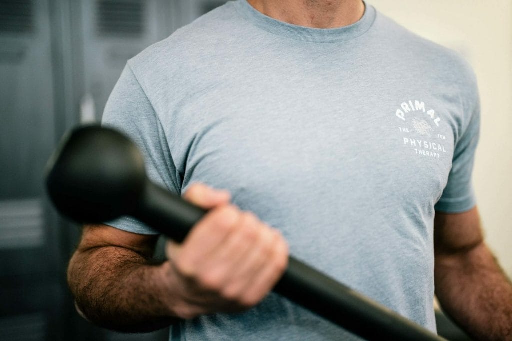 A man with a gray primal physical therapy shirt holds therapy equipment